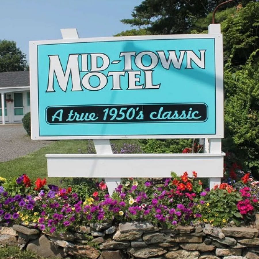 Mid-Town Motel Sign in front of hotel with teal lettering set in flower bed built up by rocks.