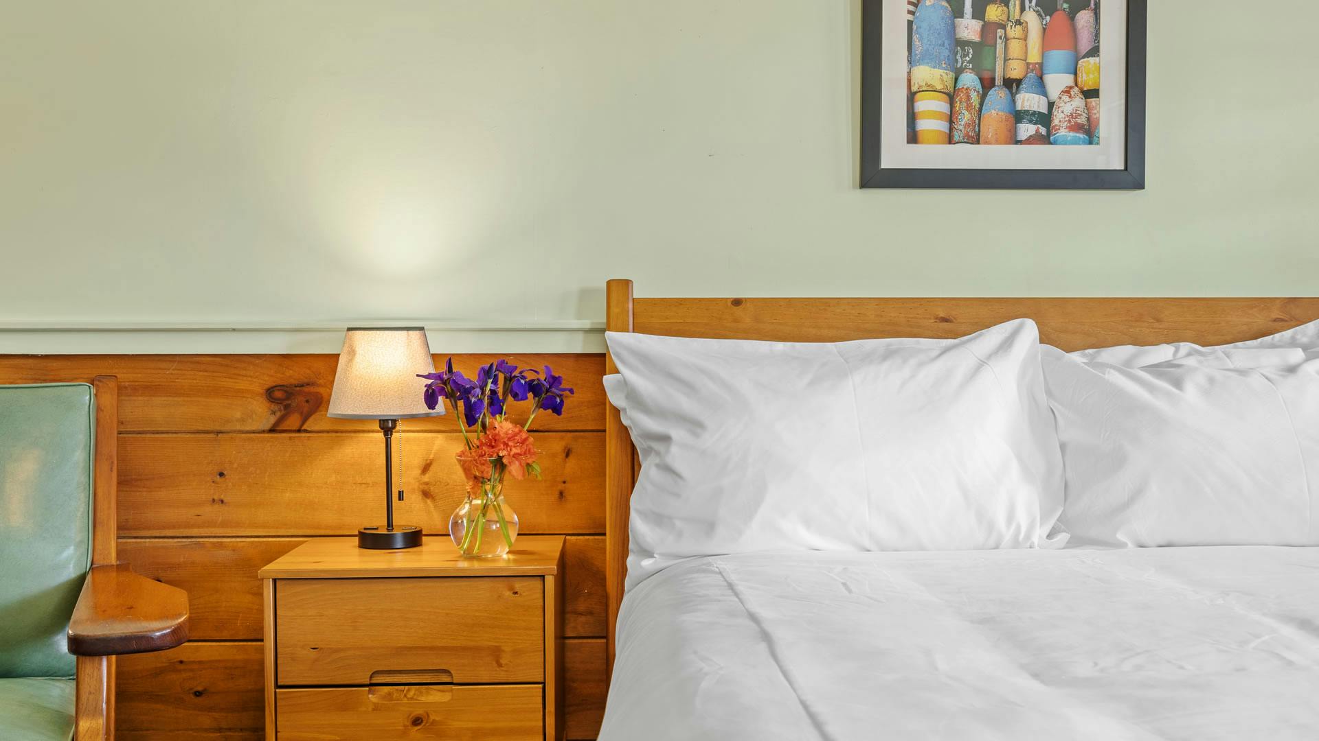 bed with white linens, flowers on nightstand, pine furniture, green vinyl chair and photo on wall of sea bobbers