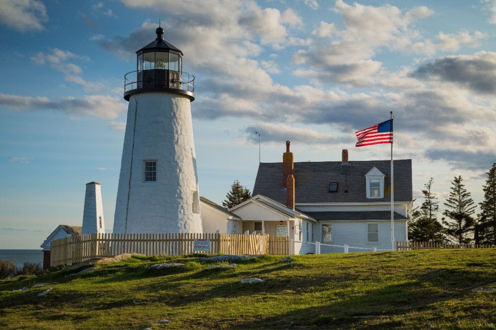 A large white lighthouse with attached building on a bluff surrounded by green grass and blue skies with clouds above