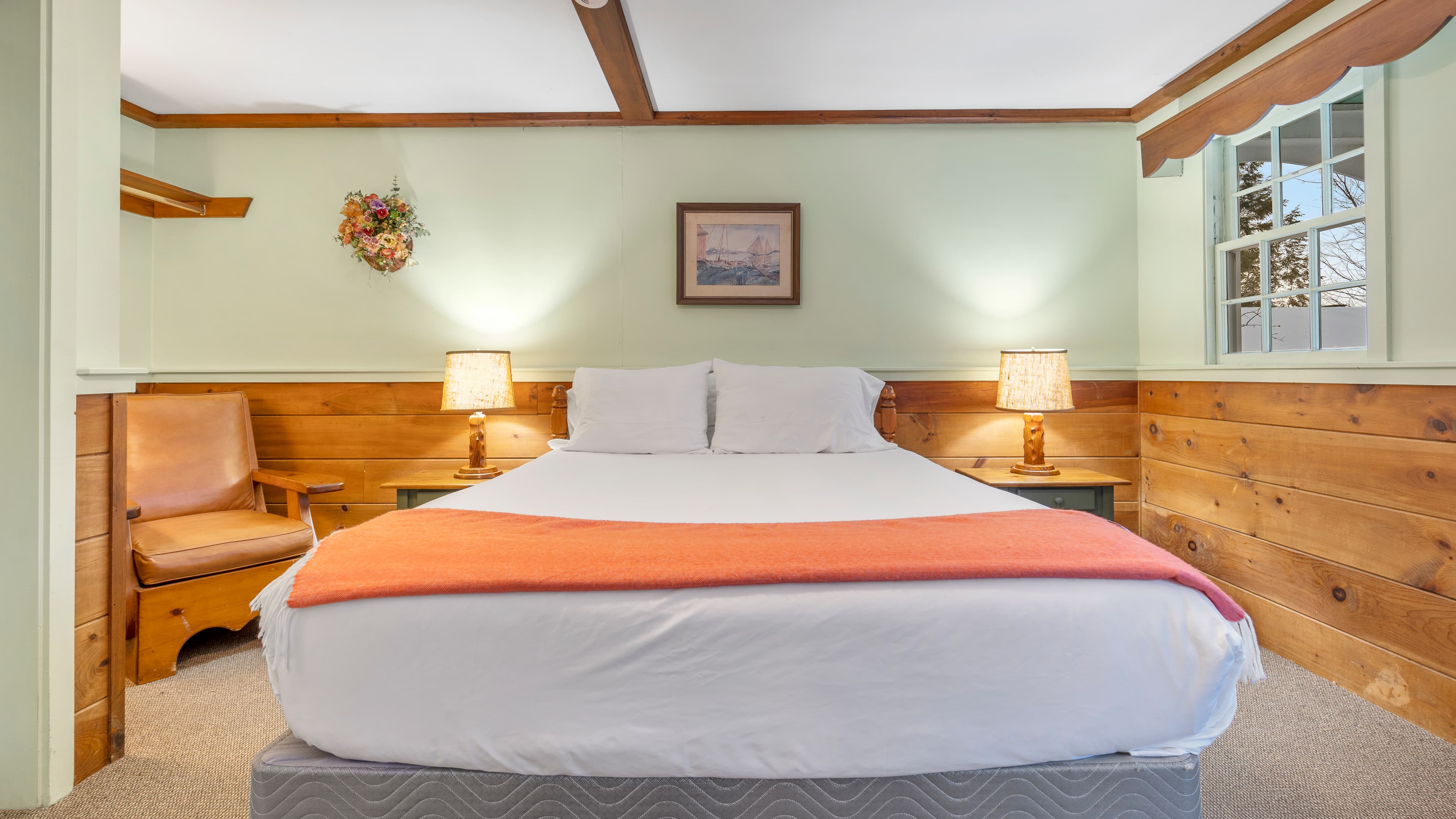 A cozy bedroom with queen bed, wood paneled walls, leather sitting chair and one window with a valence