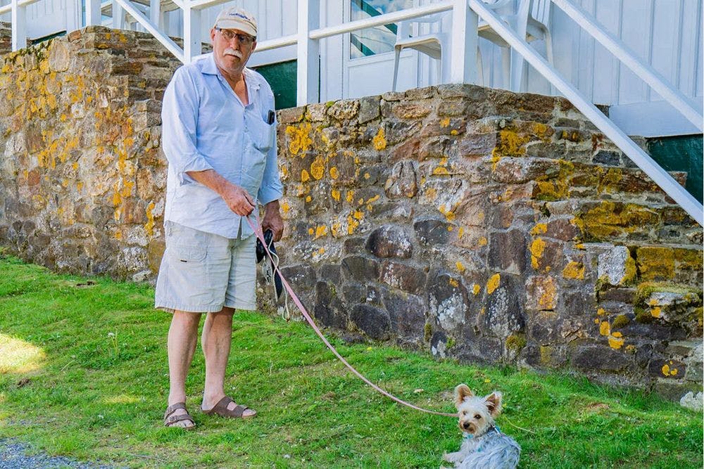 A gentleman in shorts and a blue shirt holding his dog on a leash on a grassy spot near a stone wall