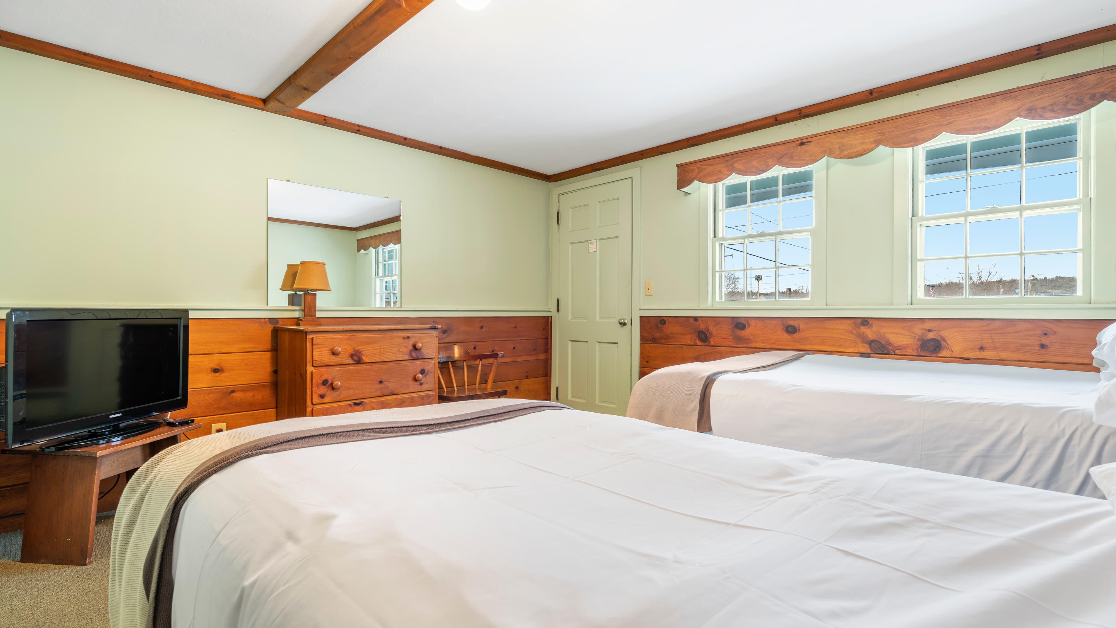 Quaint bedroom with two twin beds, dresser with a mirror, TV and two windows below a wood valence