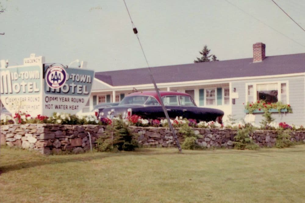 Front of a hotel with an old black and red car parked by a large sign and stone wall