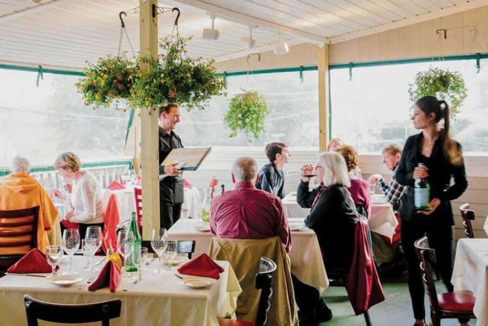 A bright and airy restaurant with walls of windows and diners sitting at tables for two being waited on by two waiters