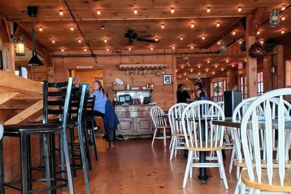 A bar and restaurant with wood paneled walls and ceiling, tables with white chairs and black bar stools