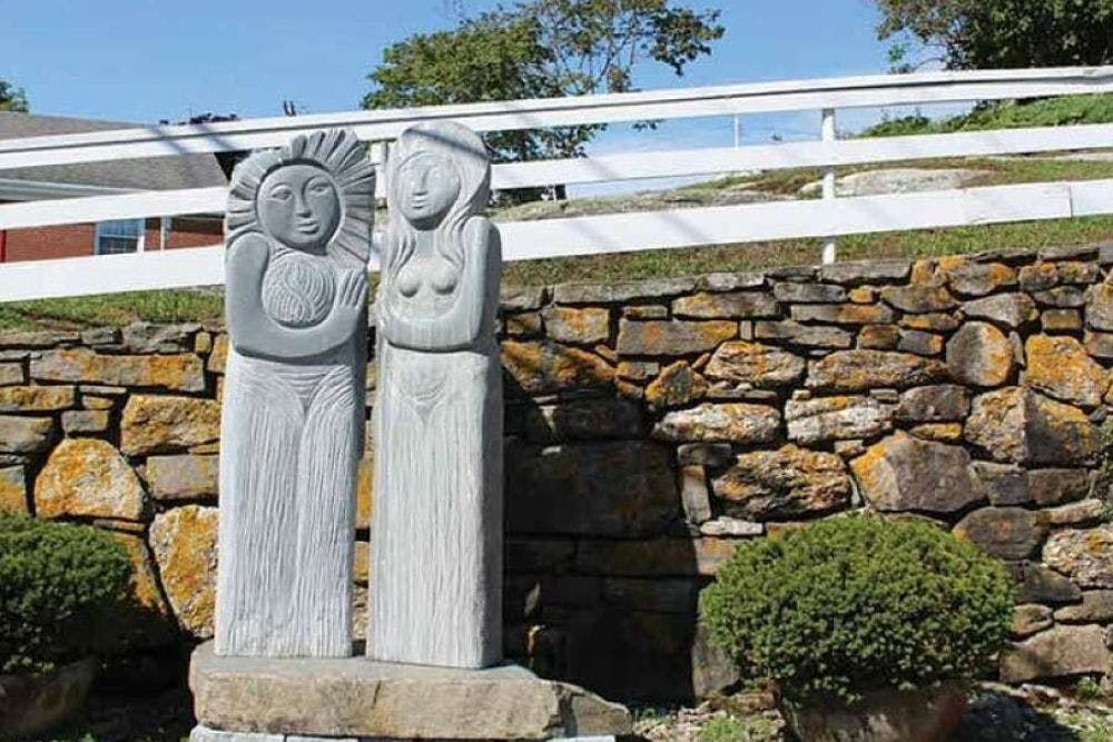 Two tall sculptures in the form of people standing side by side outside by a stone wall and white fence