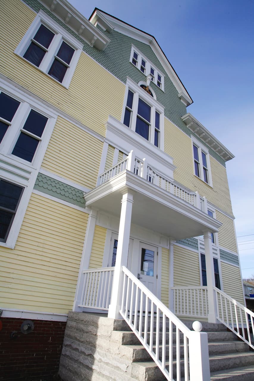A low-angle view of a yellow and green Victorian-style house with a white porch and staircase.